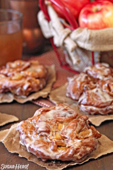 3 Apple Cider Fritters cooling on brown paper with apples in the background.