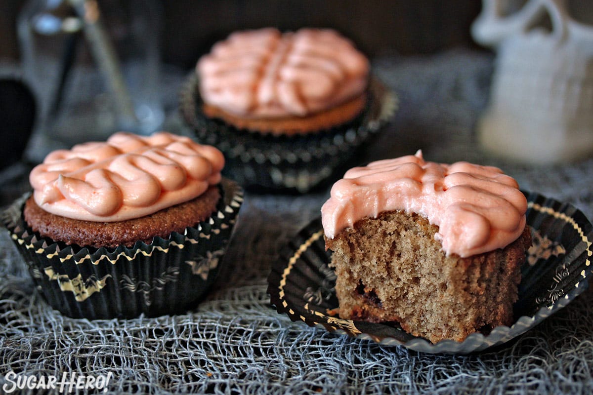3 Brain Cupcakes with bite taken out of front right cupcake on a grey webbed tablecloth.