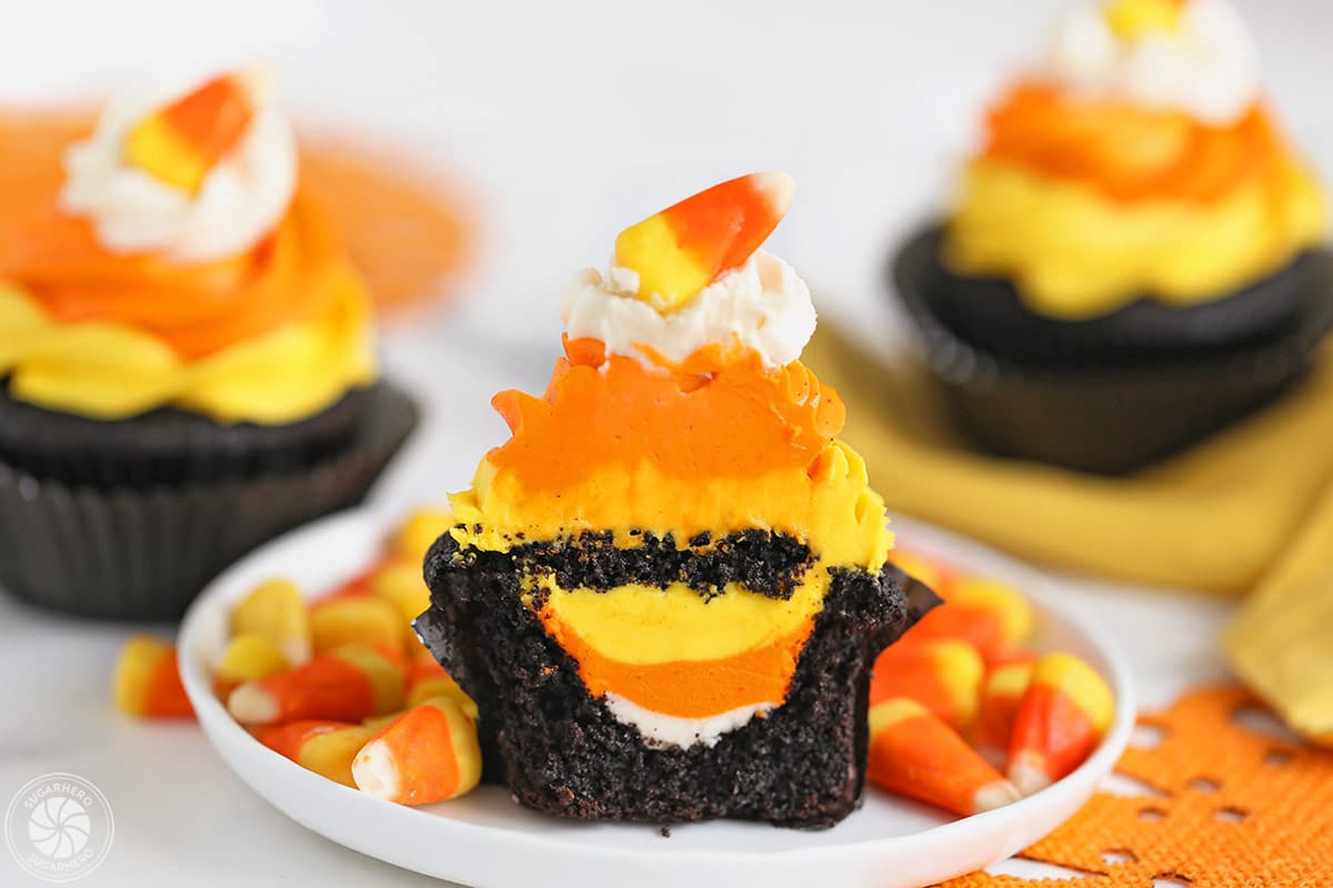 A Candy Corn Cupcake on a white plate with candy corns around it, and two cupcakes behind it.