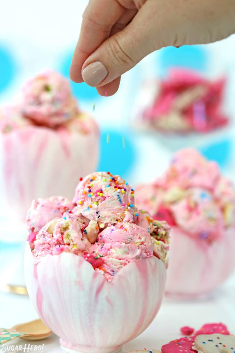 Scoops of Circus Animal No-Churn Ice Cream in a chocolate bowl with a hand sprinkling sprinkles on top.