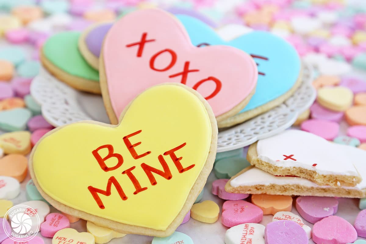 Close up of a yellow Conversation Heart Sugar Cookie with a pile of cookies on a white plate in the background and a broken cookie to the right.