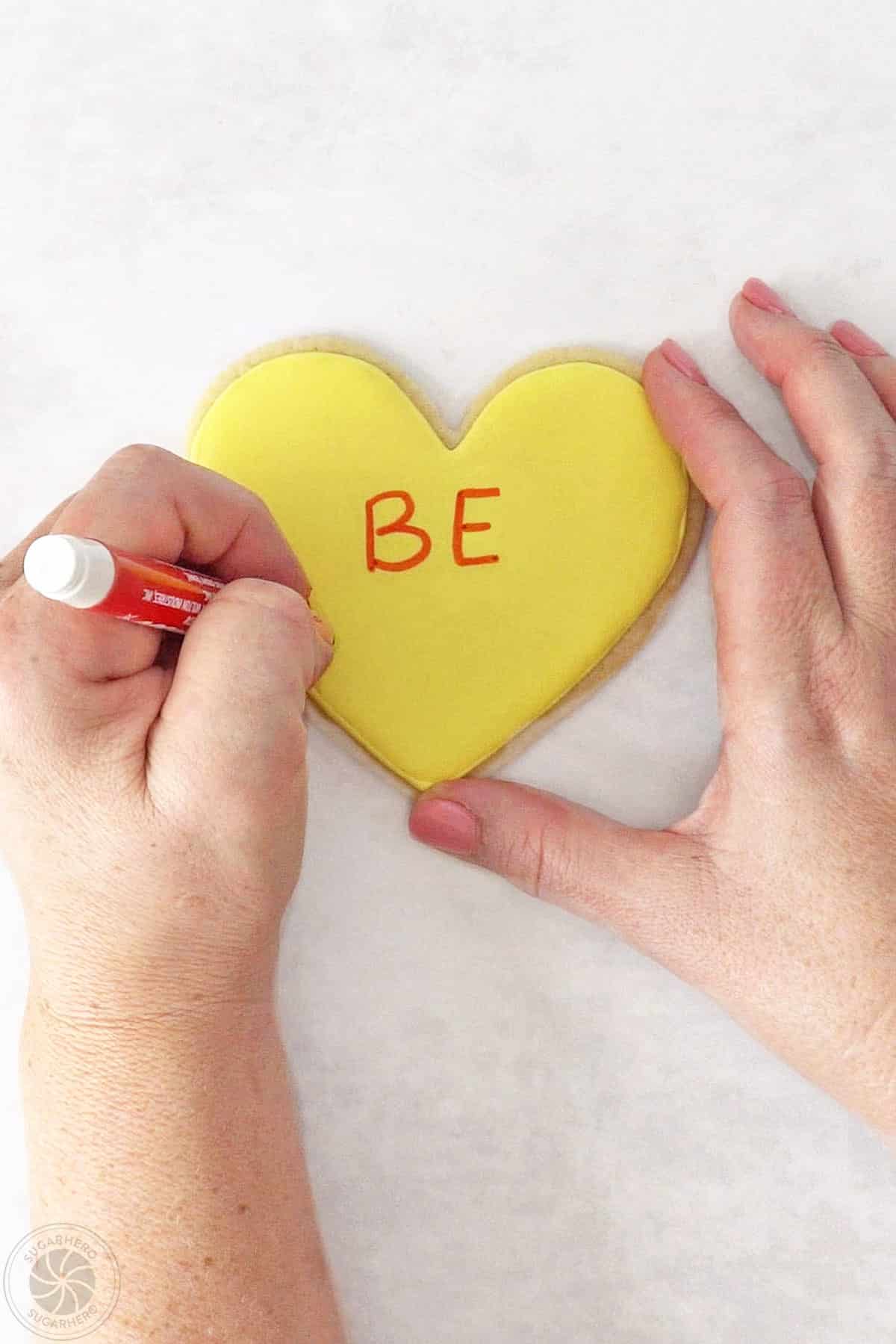 Hand using a food safe marker to write on a Conversation Heart Sugar Cookie.