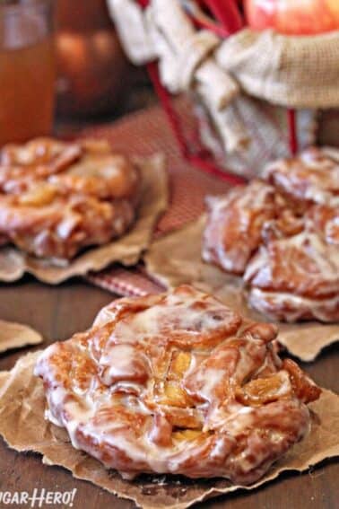 3 Apple Cider Fritters cooling on brown paper with apples in the background.