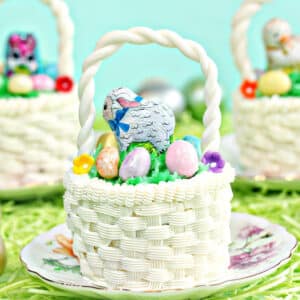 Three Easter Basket Cupcakes, each on small round plates, sitting on a bed of grass with Easter eggs scattered around.