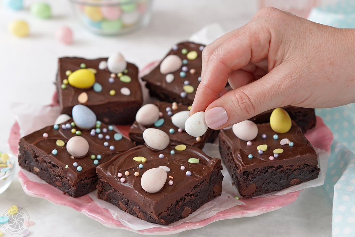 Hand with blue fingernails pressing a chocolate egg into the frosting on top of a brownie.