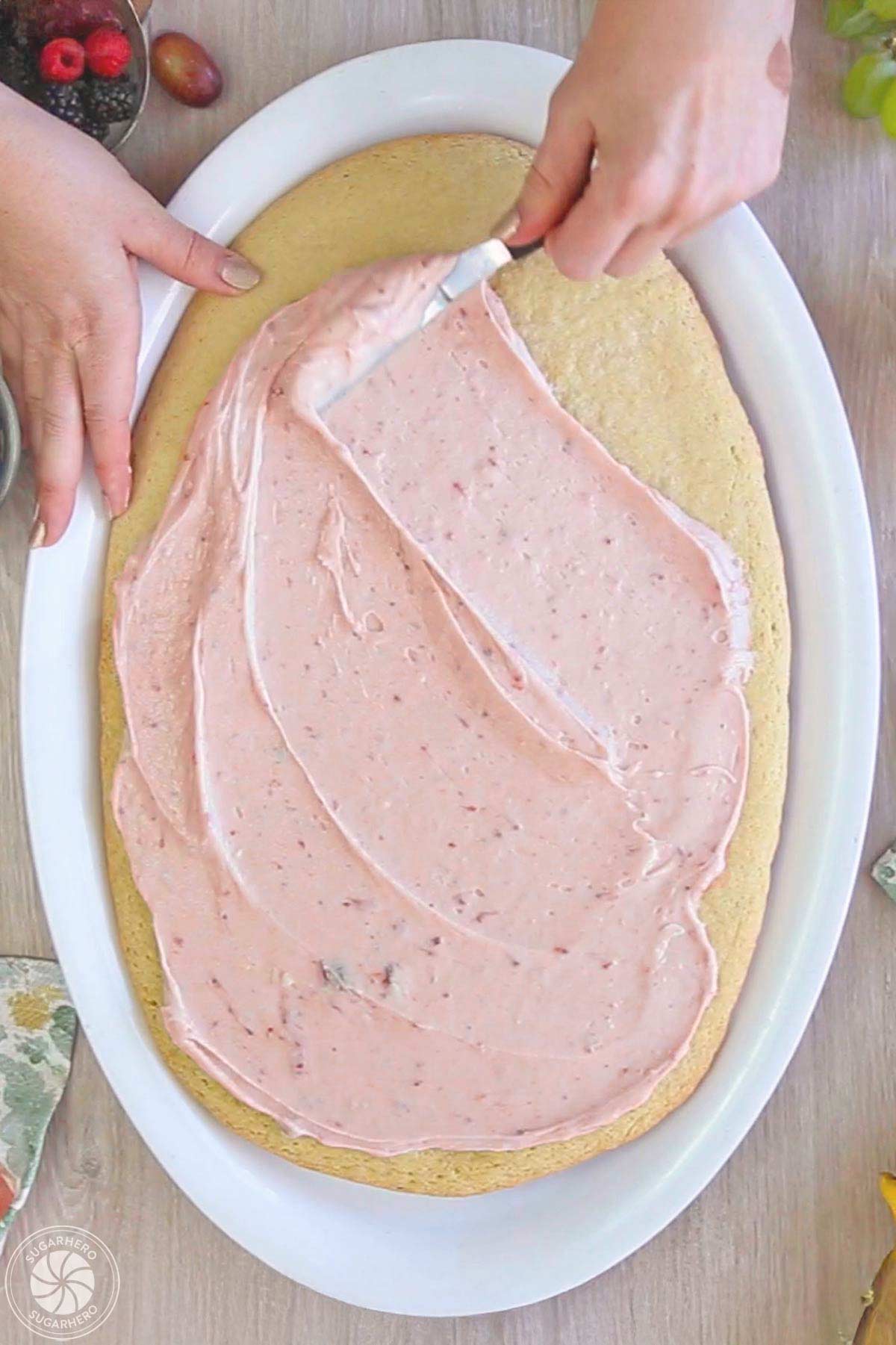 Hand spreading strawberry cream cheese frosting on top of an egg shaped sugar cookie.