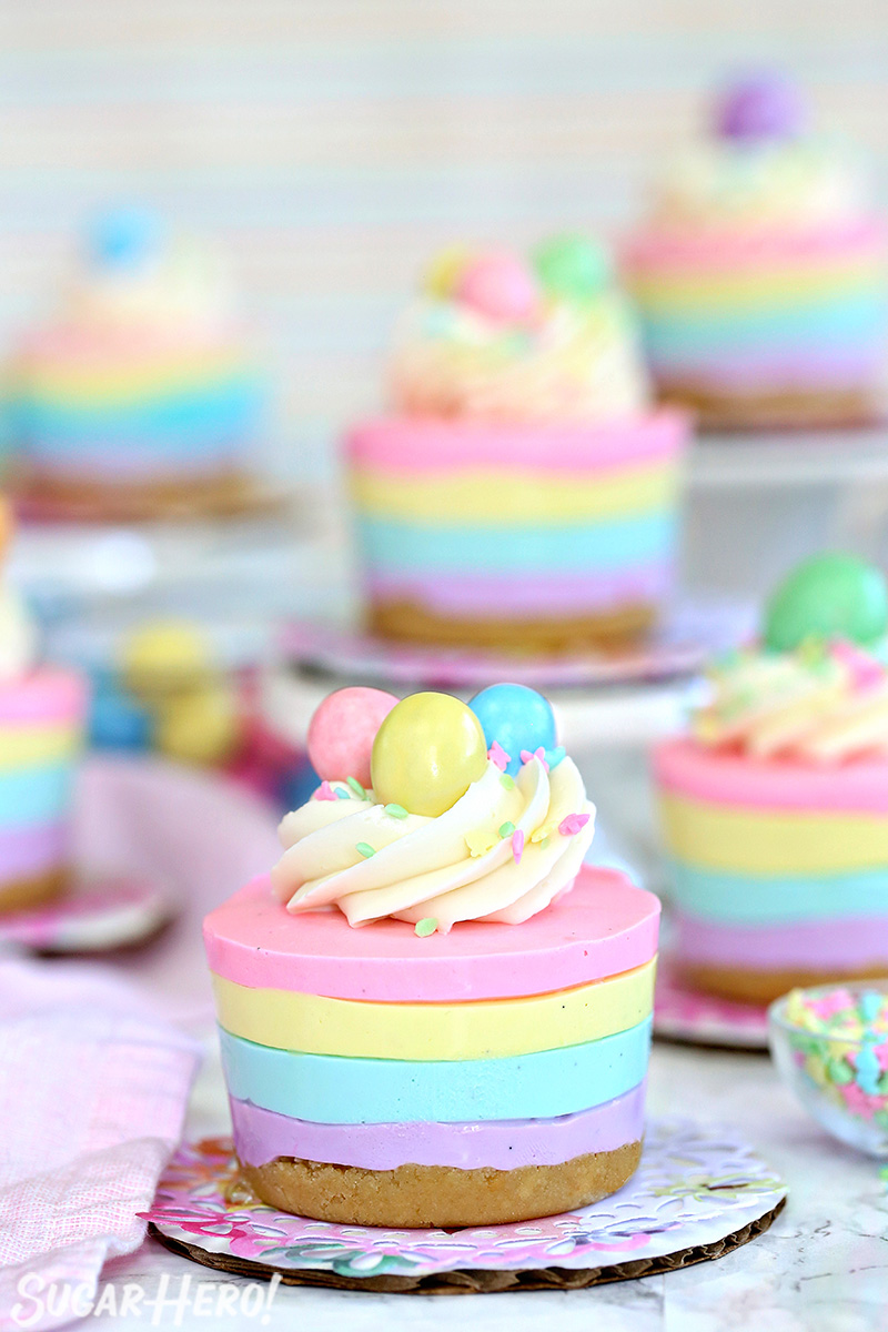 Easter no-bake mini cheesecakes topped with frosting, sprinkles, and candy eggs on colorful doilies.