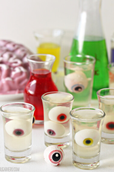 5 Floating Eyeball Jello Shots next to beakers of colorful liquid.