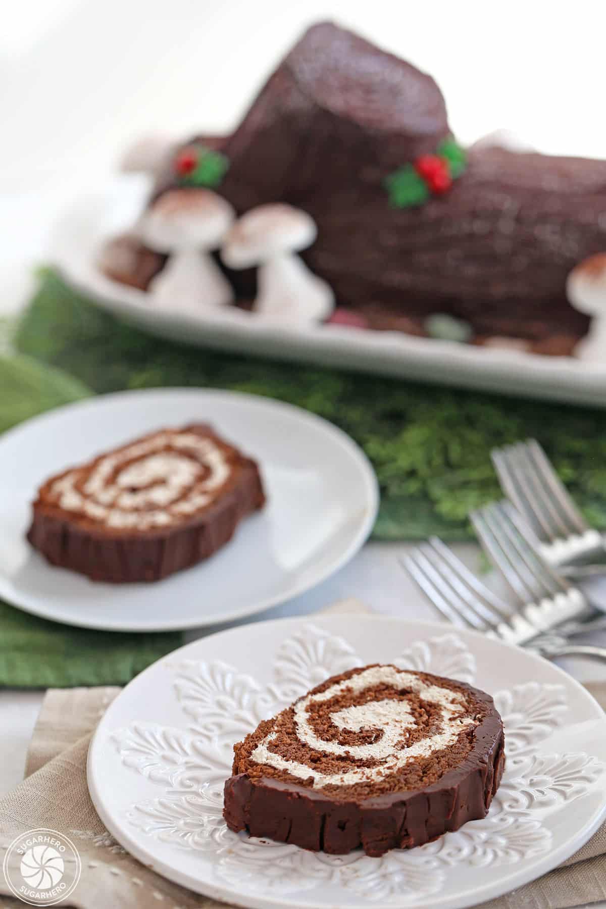 Two plated slices of Flourless Chocolate Buche de Noel with the rest of the cake in the background.