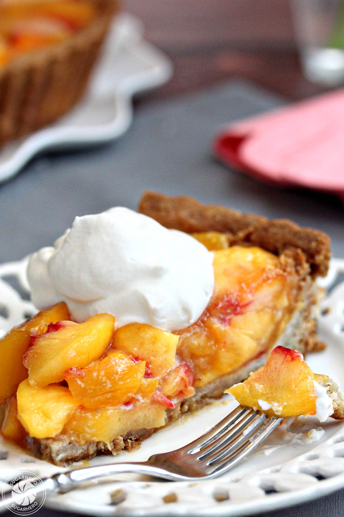 A sslice of no-bake Fresh Peach Tart, topped with whipped cream, with the whole tart in the background | From SugarHero.com