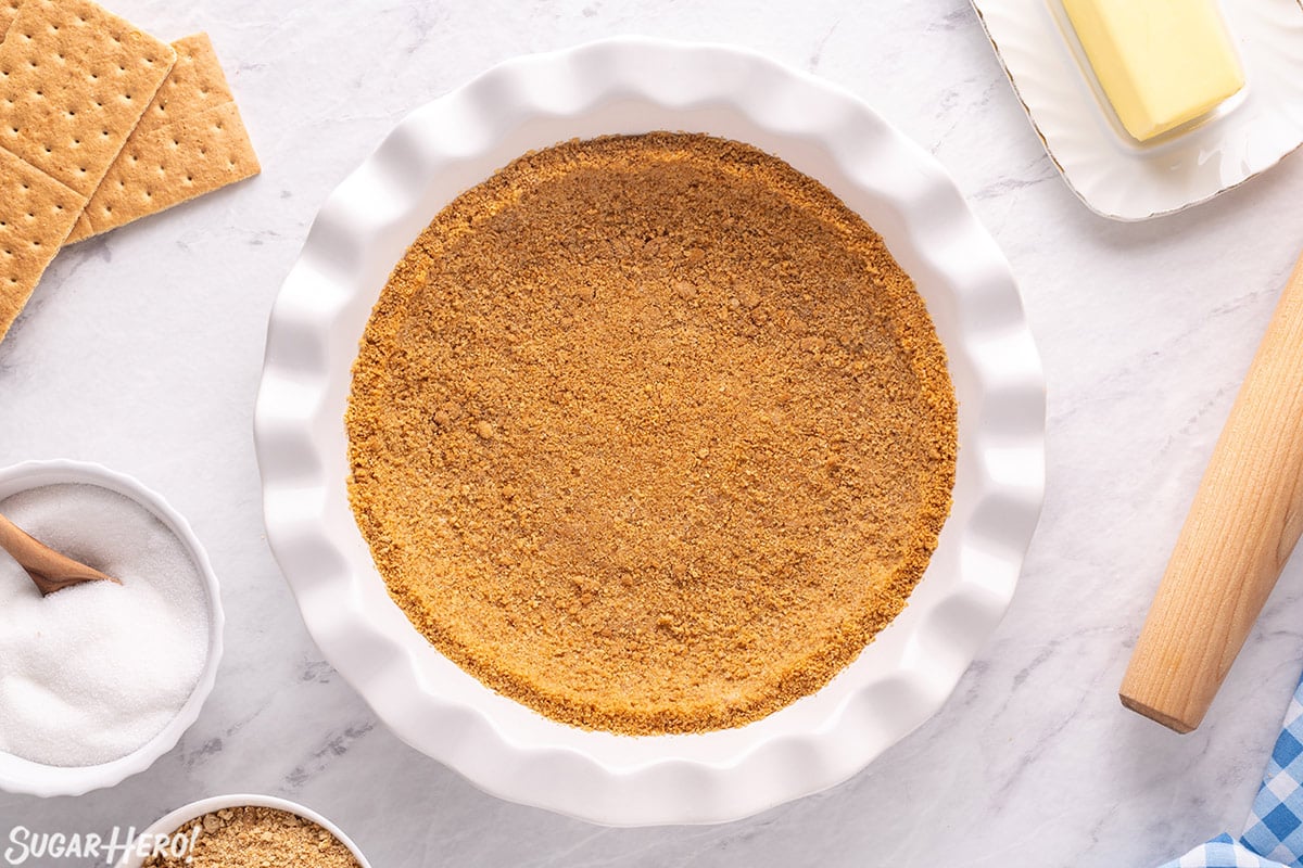Photo of graham cracker crust in a white pie pan with ingredients surrounding it.