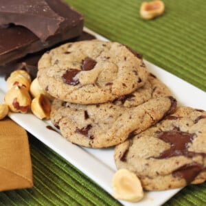 3 Hazelnut Chocolate Chunk Cookies on a white tray next to chocolate and hazelnuts.