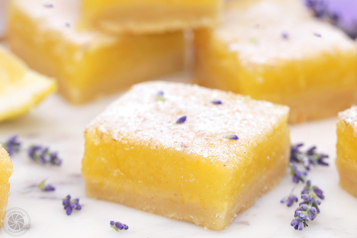 Six lavender lemon bars on a round white plate with a lavender flower on it.