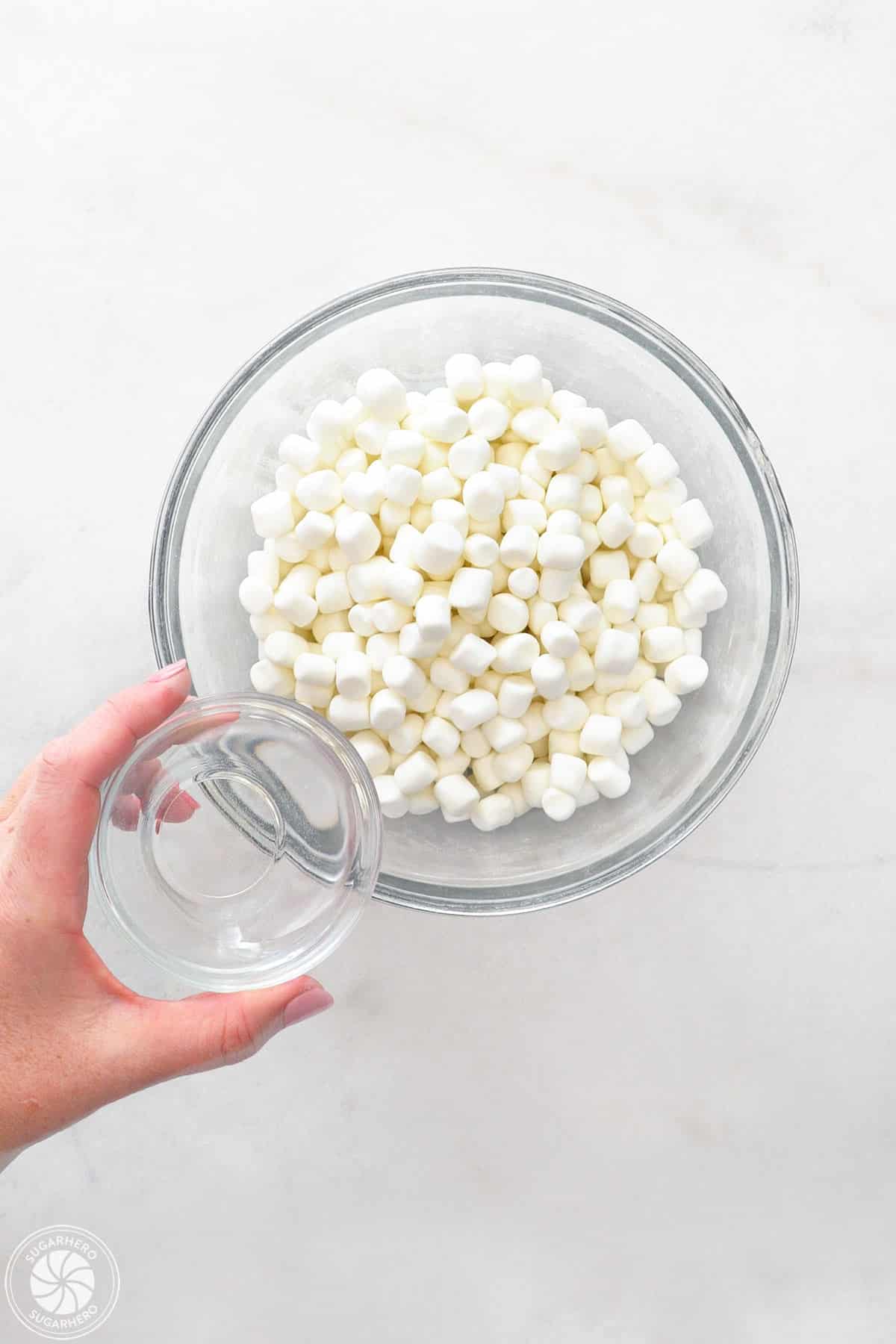 A bowl of mini marshmallows with a hand getting ready to pour in water.