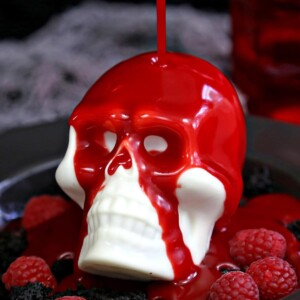 Raspberry ganache being poured over white chocolate skull on a black plate with raspberries.