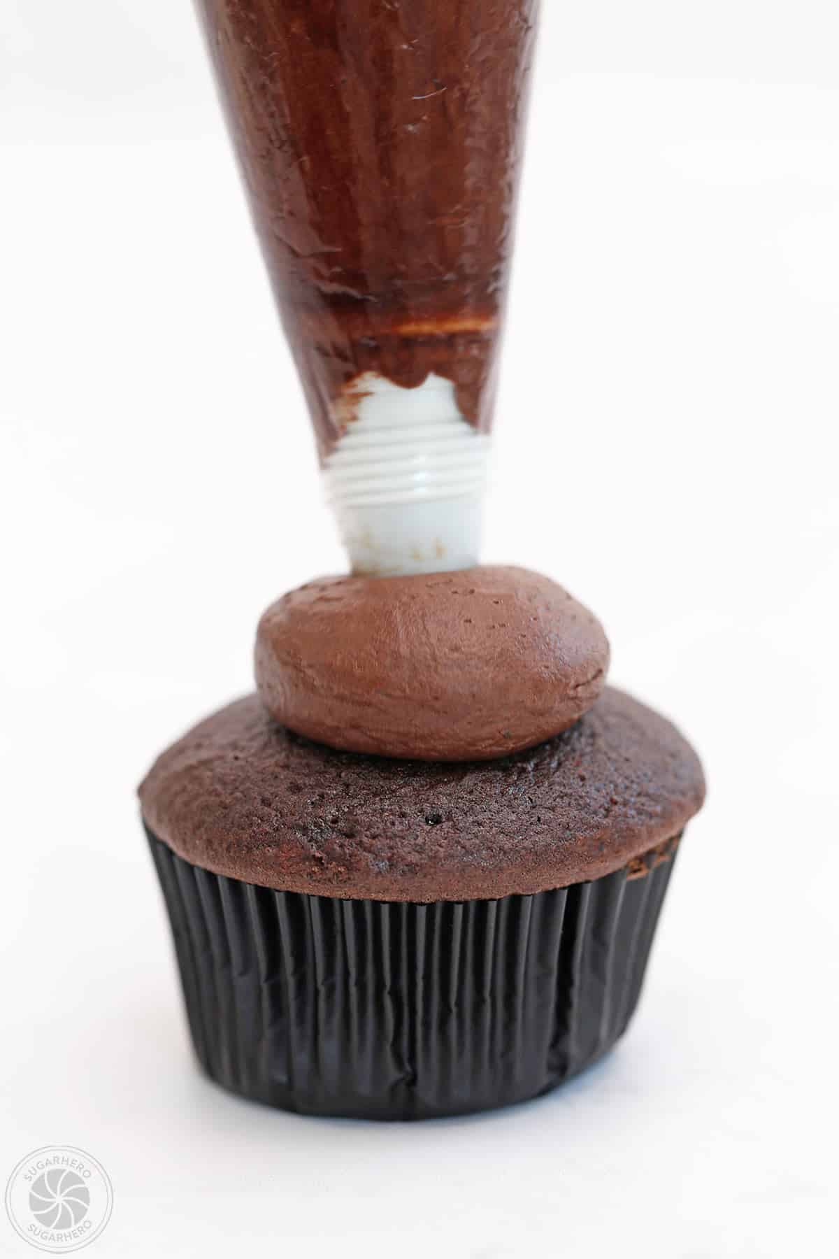 A ball of chocolate frosting being piped on top of a chocolate cupcake.