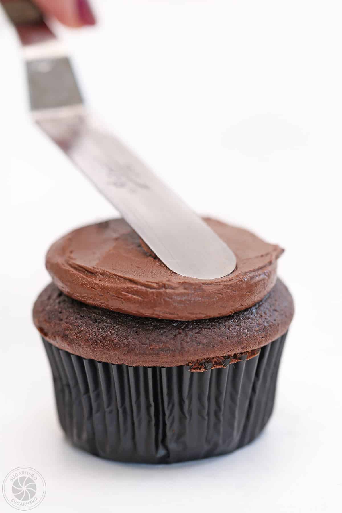 An offset spatula spreading chocolate frosting on top of a chocolate cupcake.