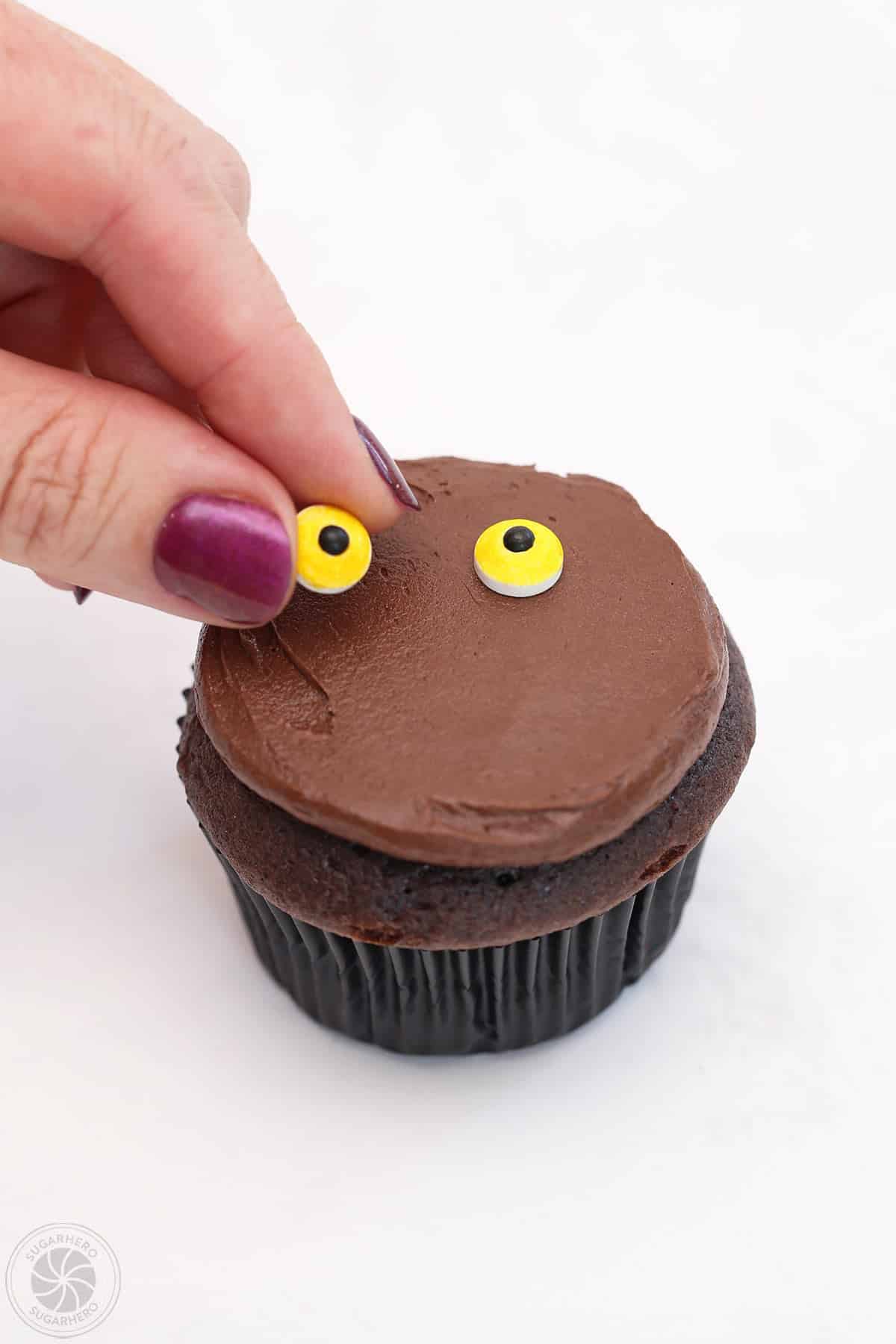 Hand placing two candy eyes on a cupcake.
