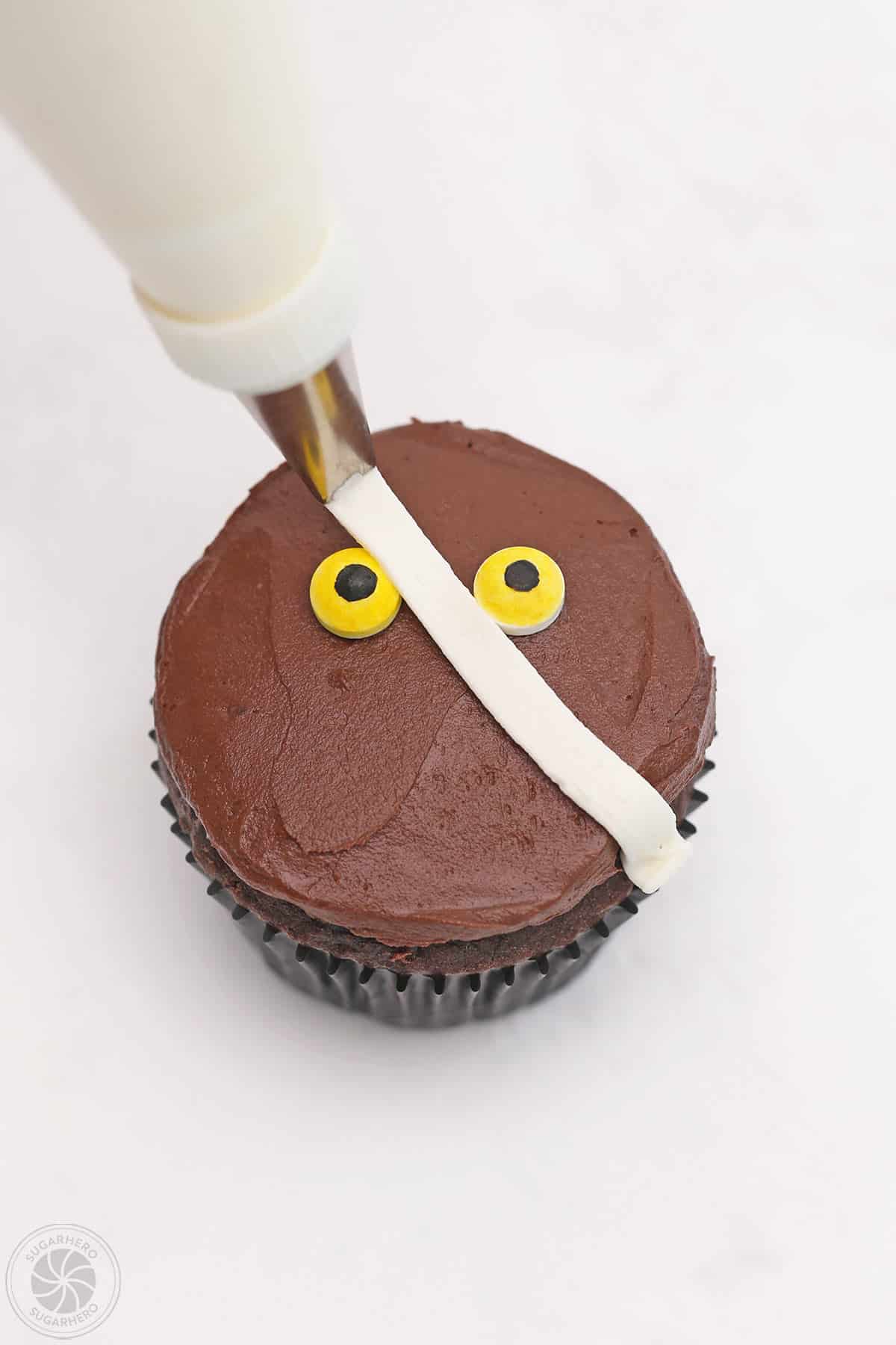 Piping a white line of frosting across the top of a mummy cupcake.