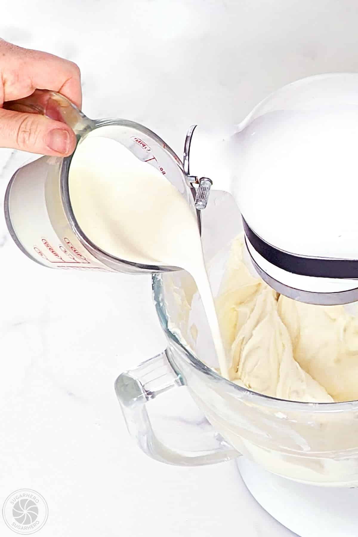 Heavy cream being poured into cheesecake batter in a mixing bowl.