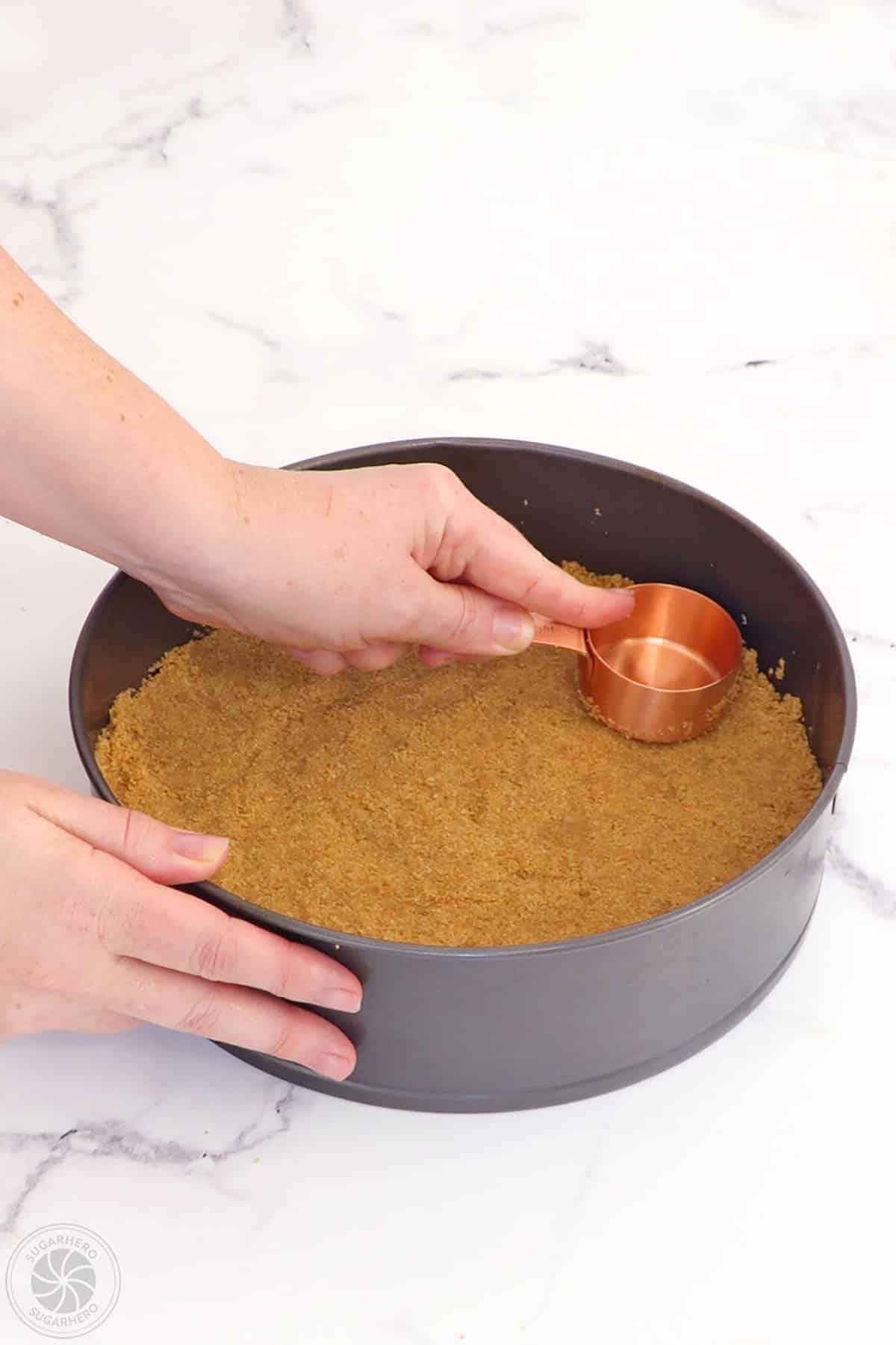 Hand pressing graham cracker crust mix into the bottom of a springform pan with a measuring cup.
