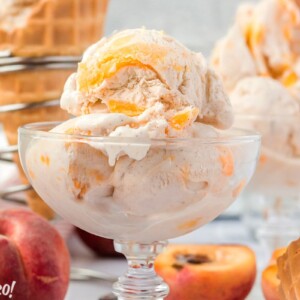 3 Scoops of No-Churn Peach Ice Cream in a clear glass bowl with peaches and waffle cones in the background.