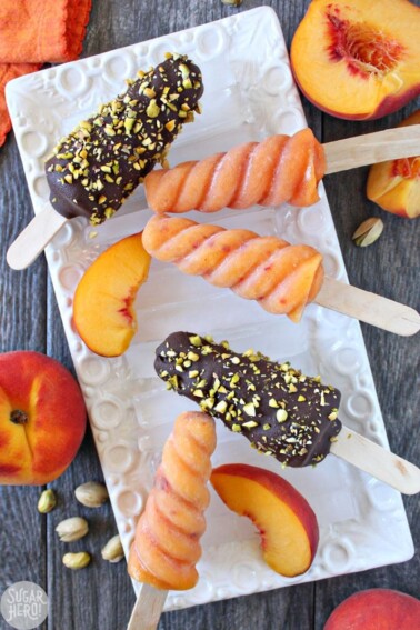An assortment of Peach Passion Pops on a white rectangular platter next to peaches and pistachios.