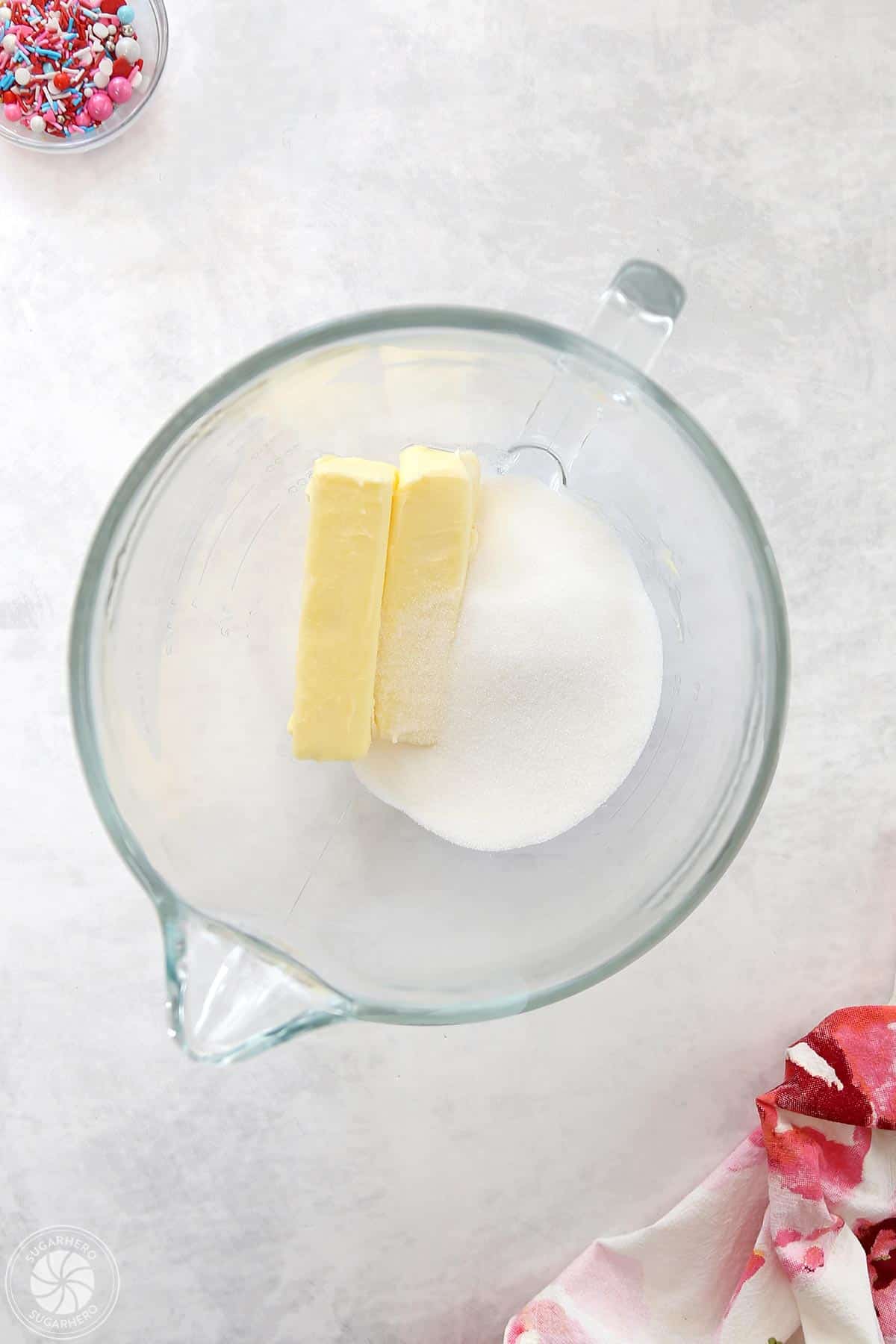 Clear glass mixing bowl with two sticks of butter and granulated sugar inside.