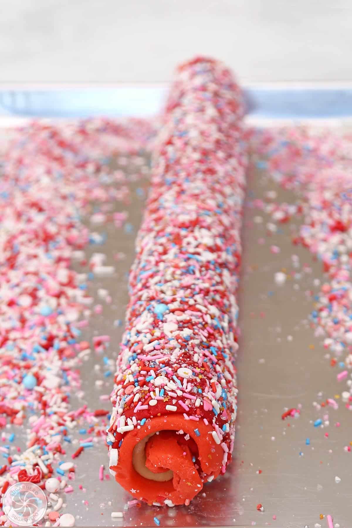 A log of Peppermint Pinwheel Cookie dough on a silver baking sheet with sprinkles scattered around.