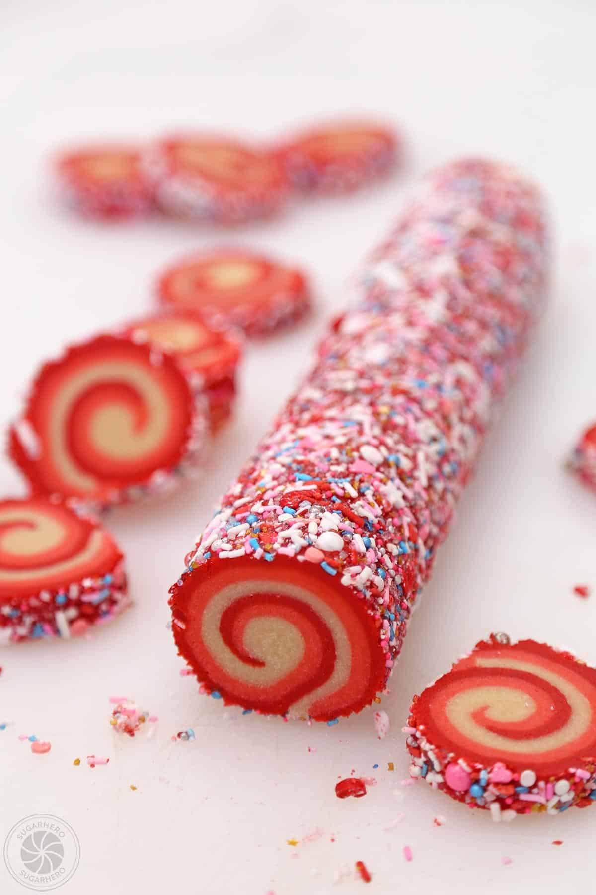 A log of Peppermint Pinwheel Cookie dough, rolled in sprinkles, with sliced cookies scattered around it.