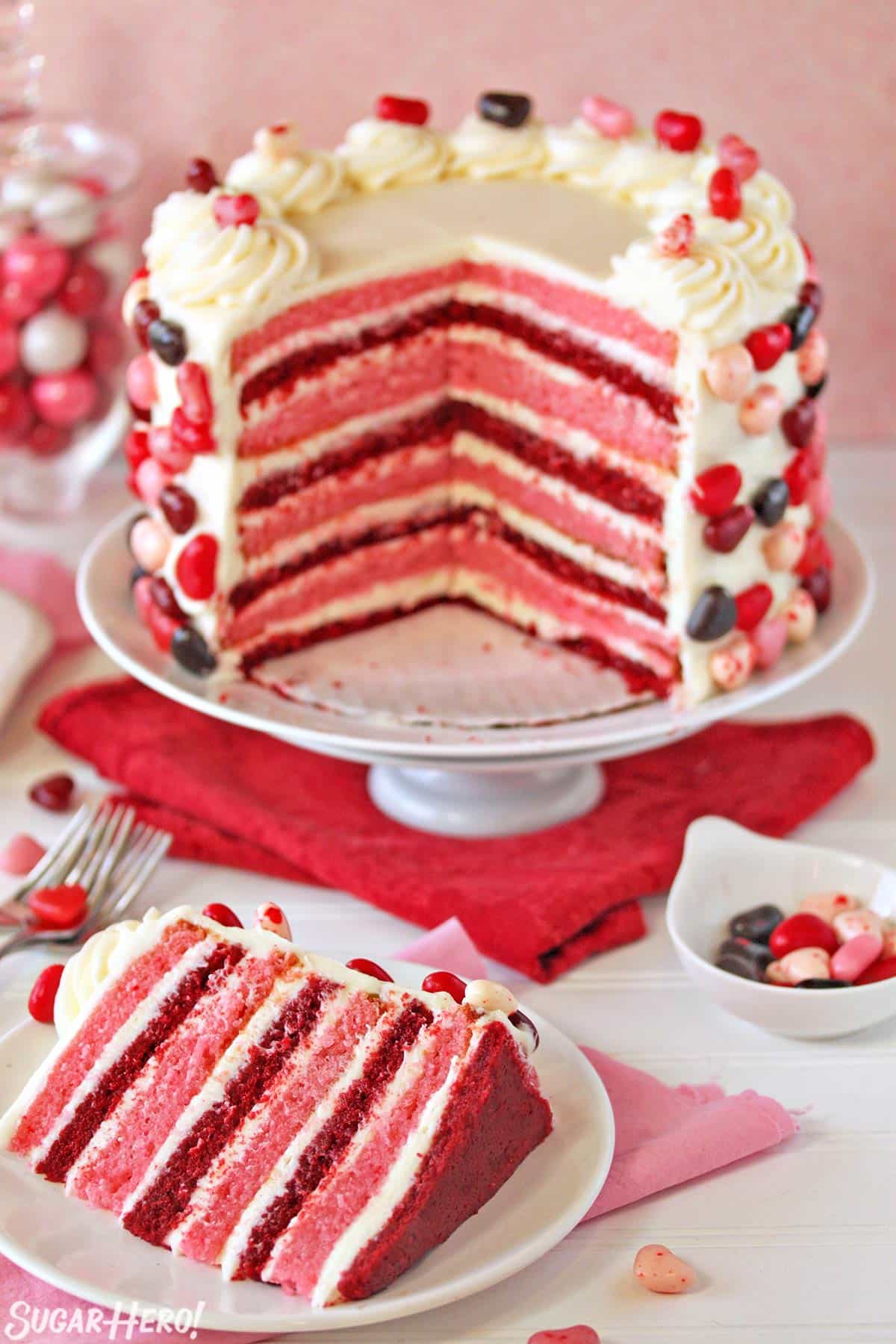 Pink and red velvet layer cake with several slices taken out, and a slice of cake displayed in front on a white plate.