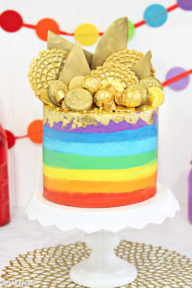 Gold-Topped Rainbow Cake on a white cake stand in front of colorful bunting in the background.