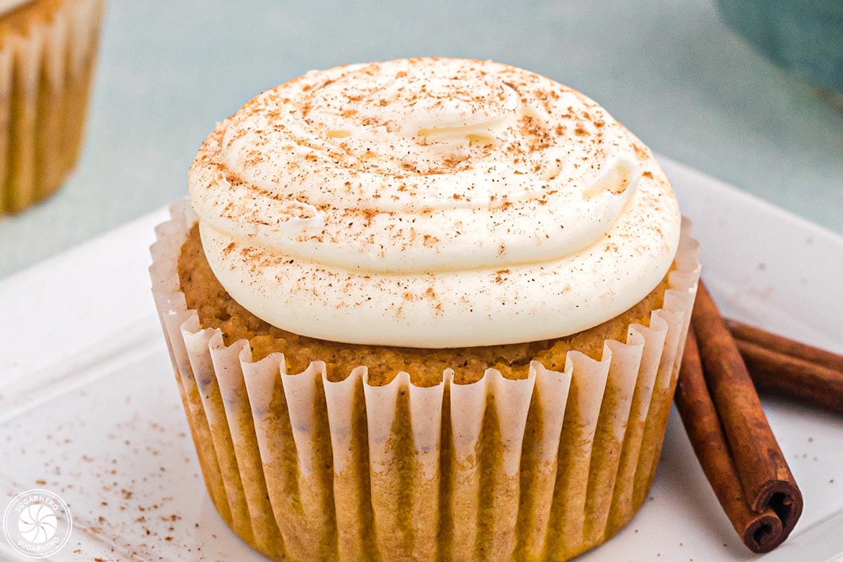 Pumpkin spipce cupcake with cinnamon sticks beside it sitting on two stacked white square plates.