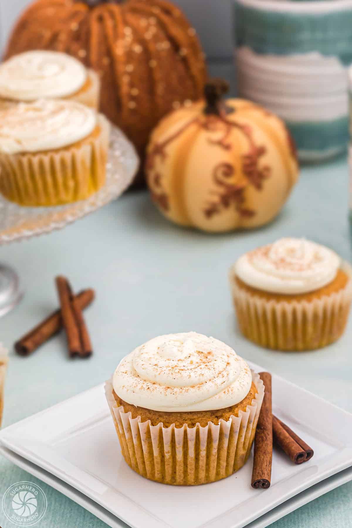 Portrait style photo of pumpkin spice cupcake on white square plate with other cupcakes and fall decor in the background.