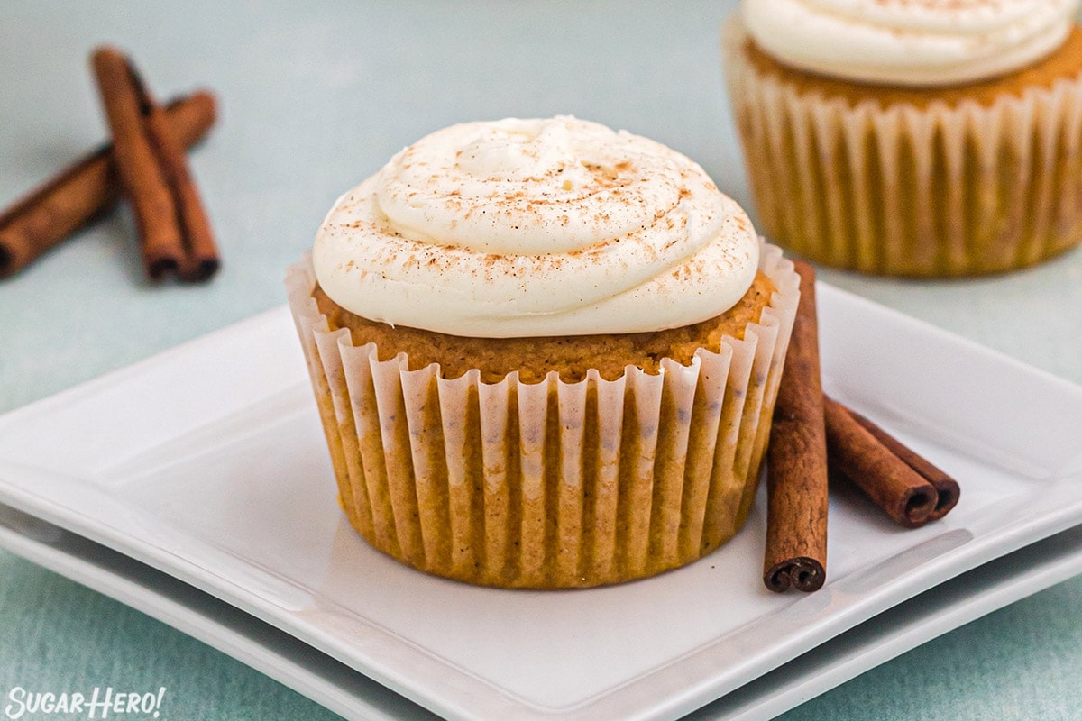 Pumpkin spice cupcake sitting on two stacked square white plates with two cinnamon sticks to the right of it.
