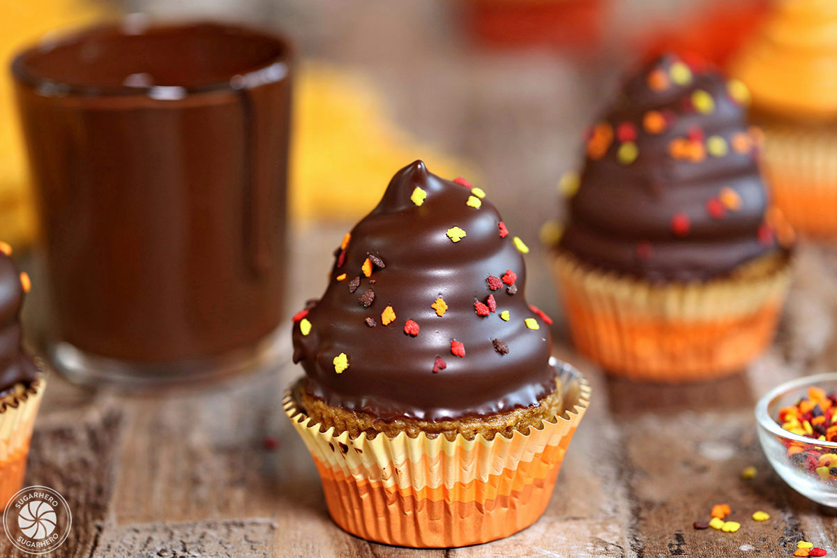 2 Pumpkin Spice Hi-Hat Cupcakes on a wooden surface next to a dripping cup of chocolate glaze and sprinkles.
