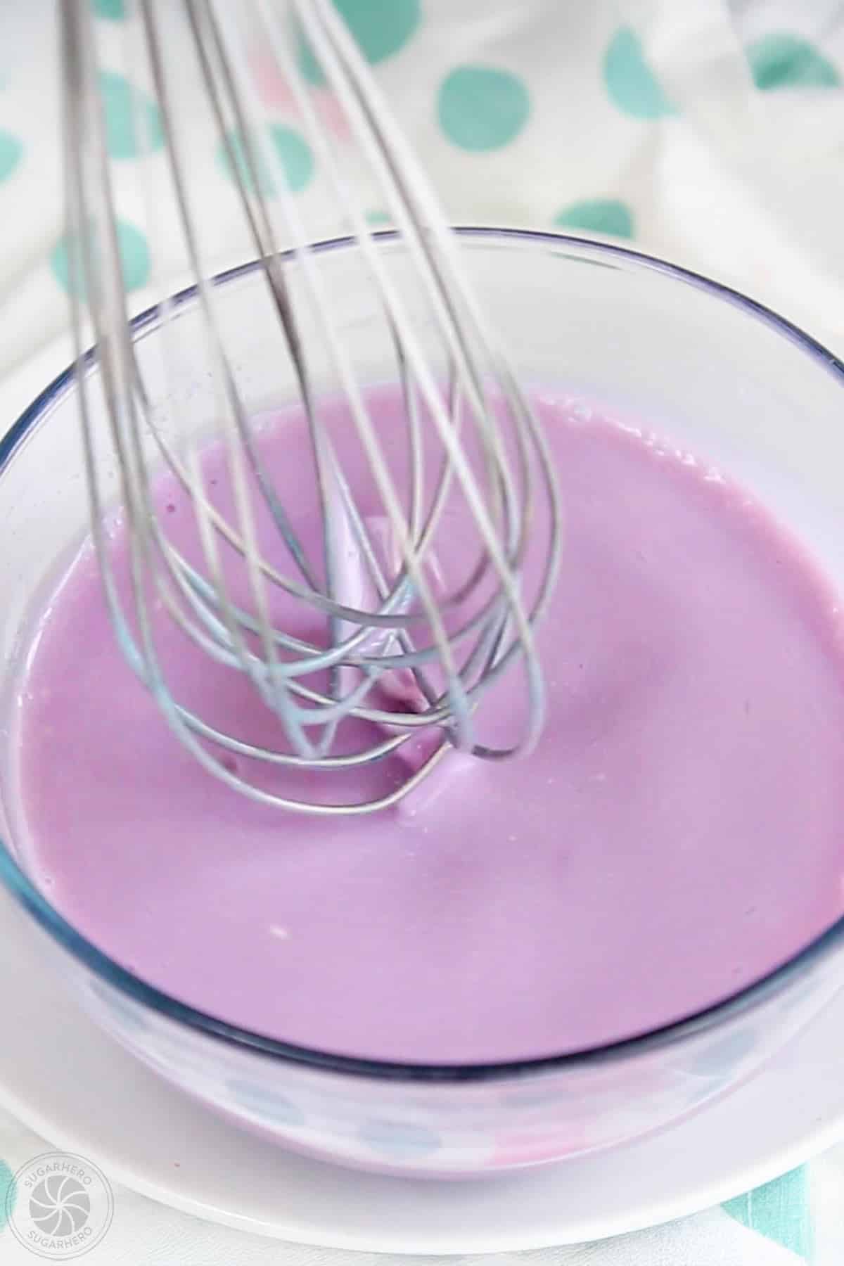 A mixing bowl of creamy lavender gelatin with a whisk above it.