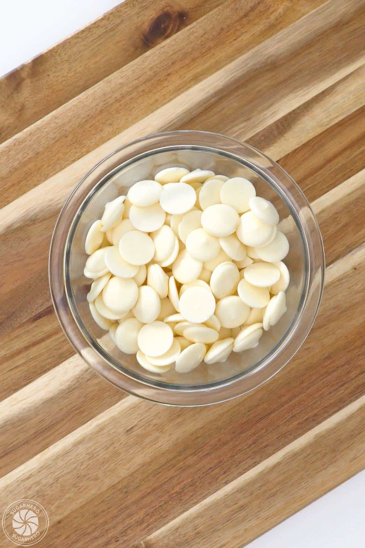 Glass bowl of white candy coating wafers on a wooden cutting board.