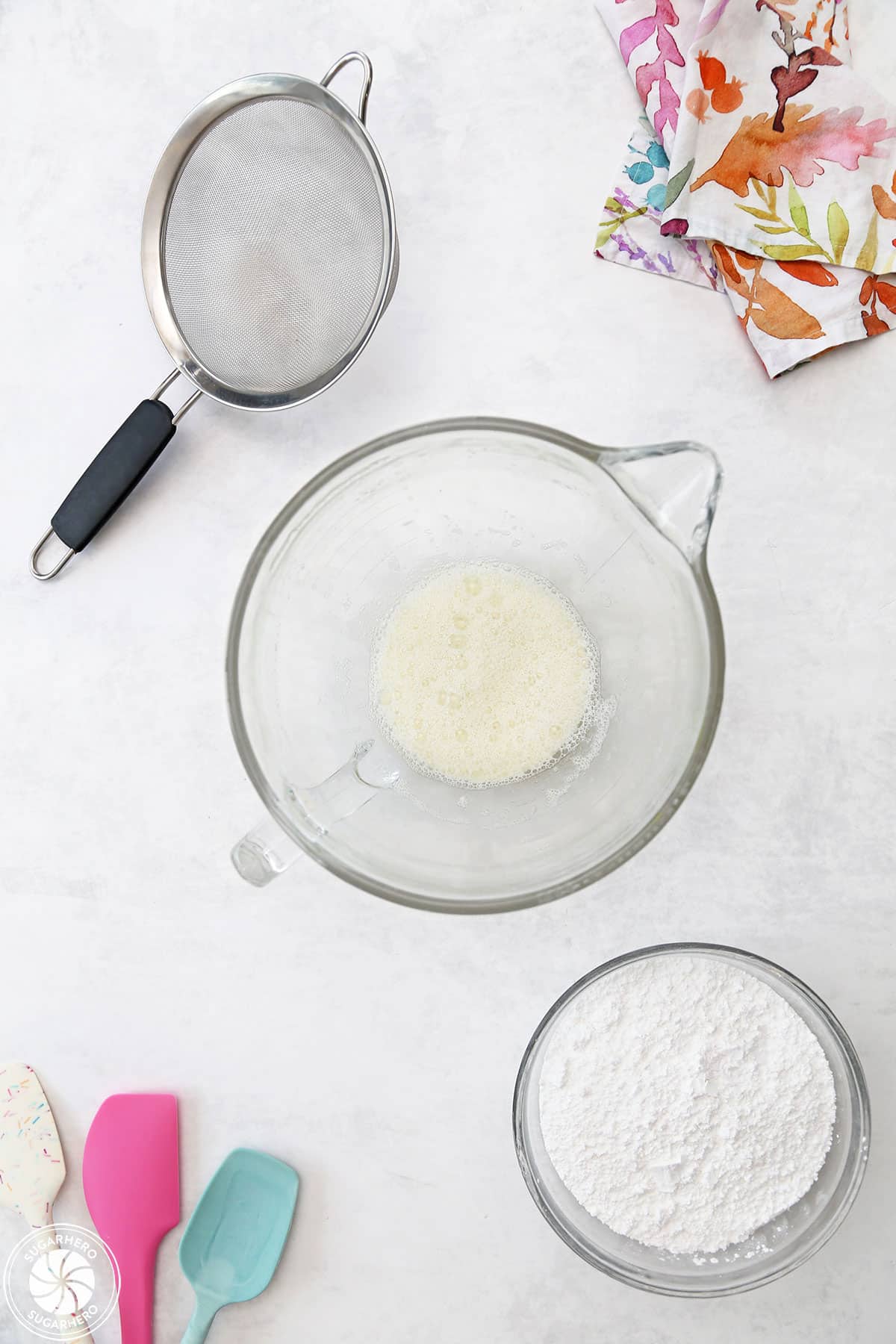 Meringue powder in a mixing bowl, with a bowl of powdered sugar and a strainer nearby.