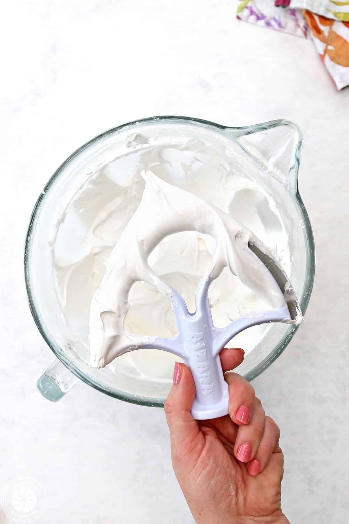 Hand holding a paddle with stiff royal icing on it, over a mixing bowl full of royal icing.