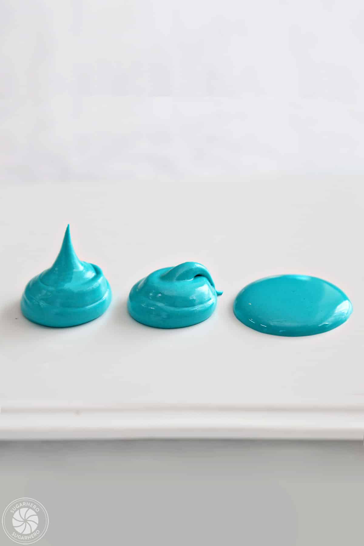 Three dollops of teal royal icing on a white plate, showing different consistencies of the icing.