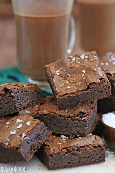 A pile of Salted Fudge Brownies cut into squares next to a bowl of salt a glass of hot chocolate.
