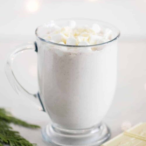 Side view of a clear mug of Snickerdoodle White Hot Chocolate next to a piece of white chocolate and cedar.
