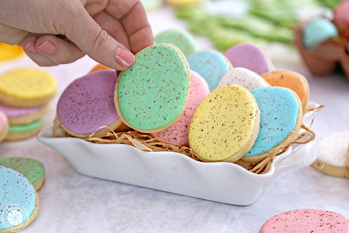 Speckled Easter Egg Sugar Cookies in a white container, with a hand reaching in and grabbing a green cookie.