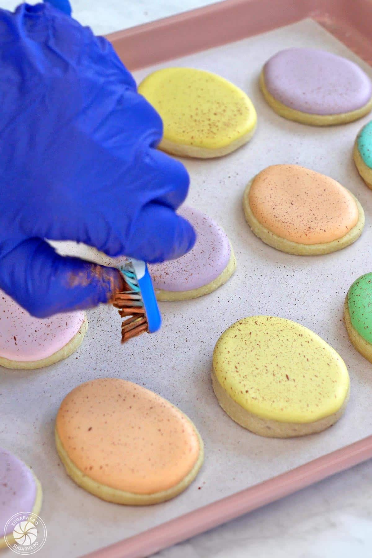Gloved hand using a toothbrush to speckle cocoa powder over egg cookies.
