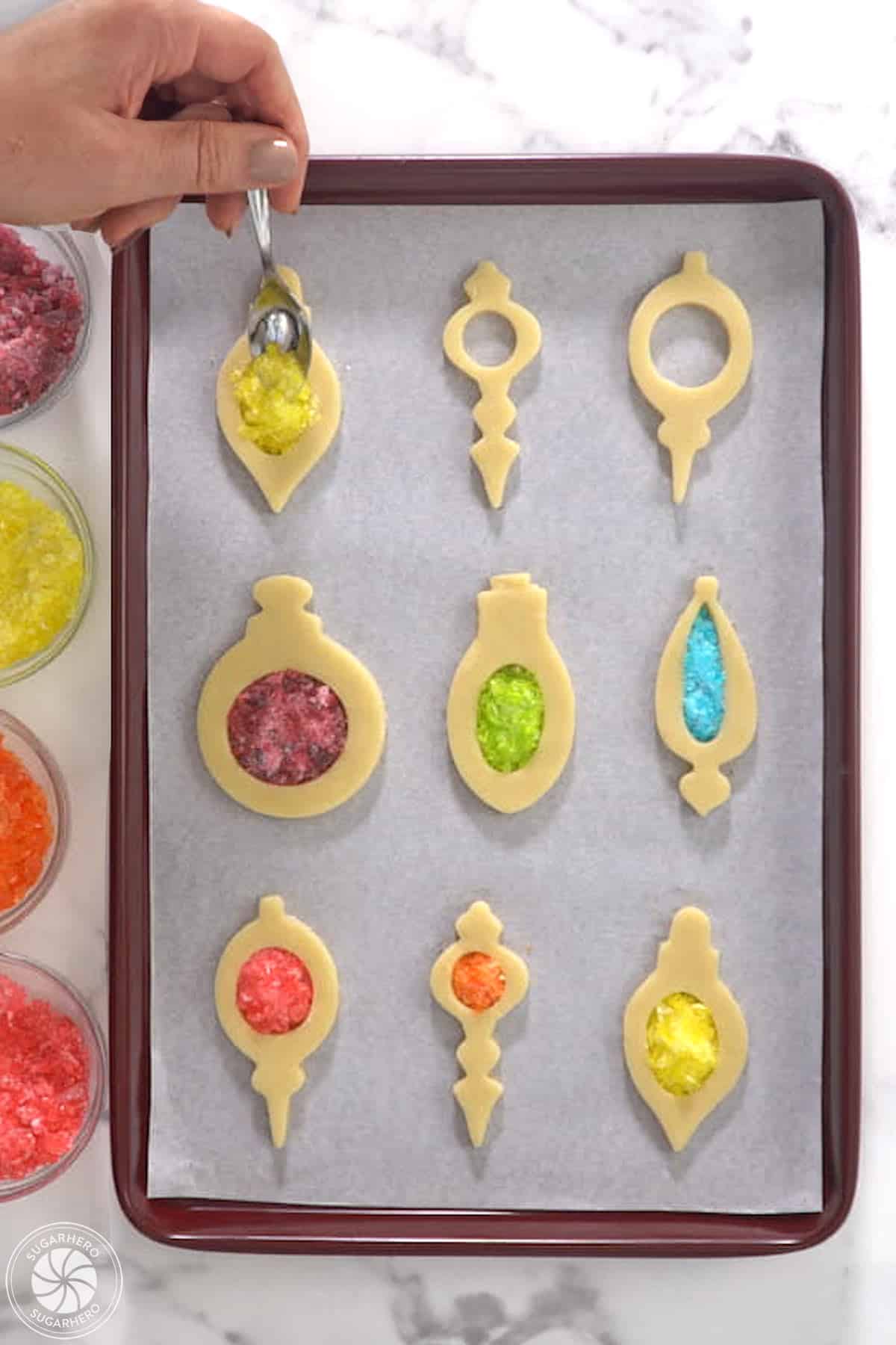Cut out cookie shapes on a baking tray with a hand filling the centers with crushed candy.