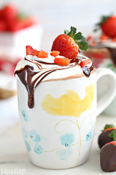 Strawberry hot chocolate in a floral mug with whipped cream and sliced strawberries on top next to chocolate covered strawberries.