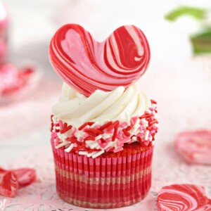 Close-up of cupcake with white buttercream, pink and red sprinkles, and a large chocolate heart on top.