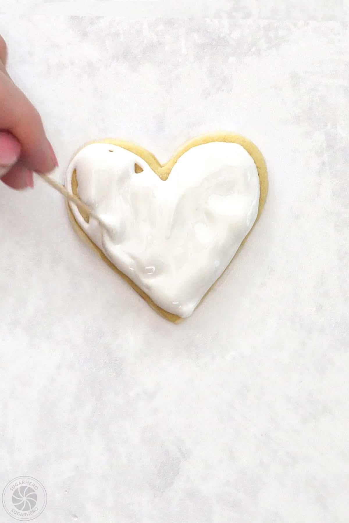 Hand using a toothpick to spread the royal icing into the center of a Valentine's Day Sugar Cookie.