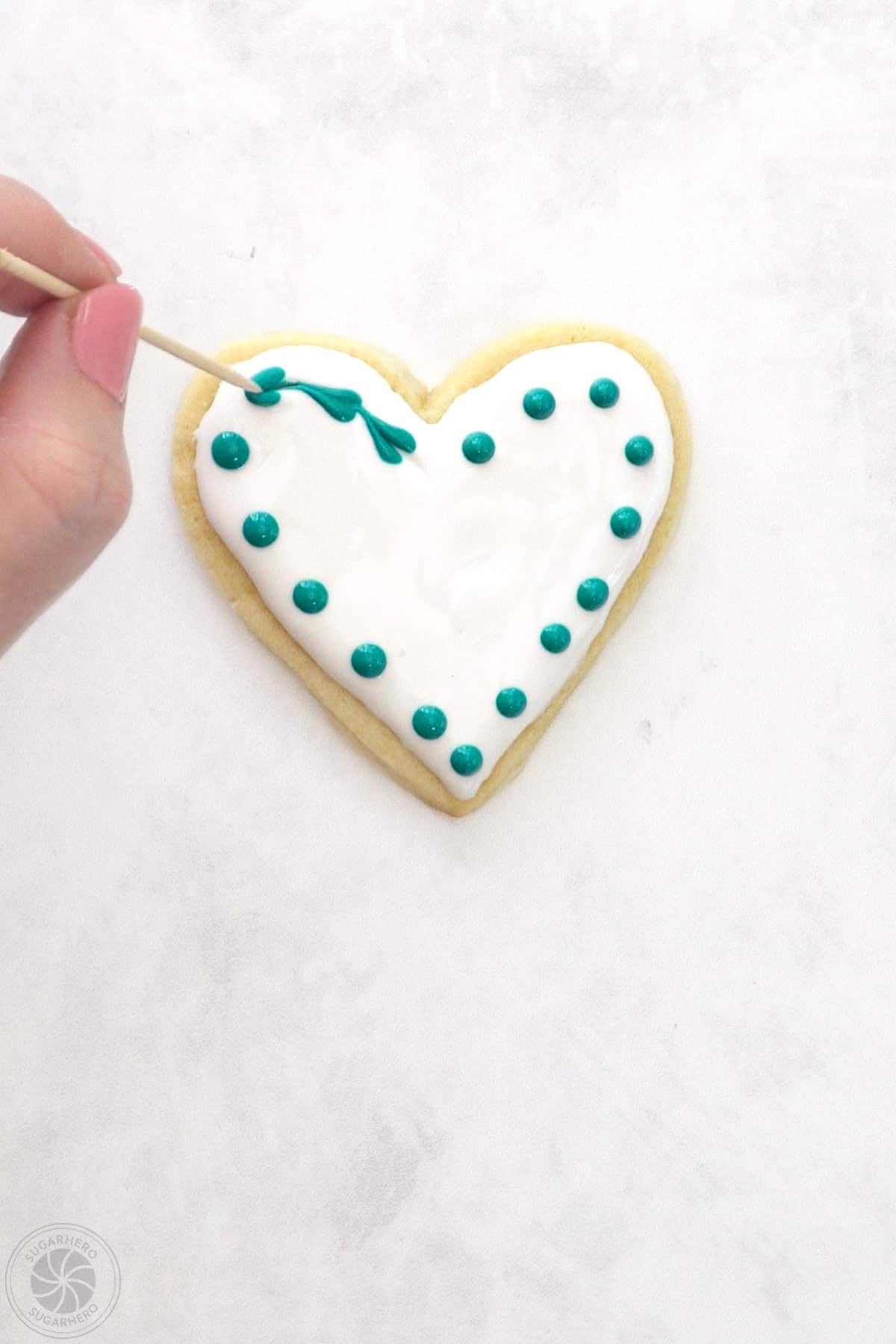 Hand using a toothpick to go through the center of dots outlining the outside of a Valentine's Day Sugar Cookie.